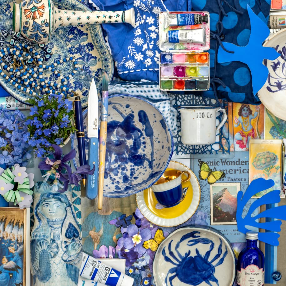 A colorful flat lay of blue-themed objects includes ceramics, flowers, watercolor paints, brushes, books, jewelry, and decorative illustrations arranged closely together.
