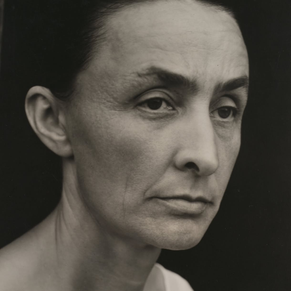 A black-and-white close-up portrait of Georgia O'Keefe with dark hair pulled back, looking slightly to the side against a dark background.