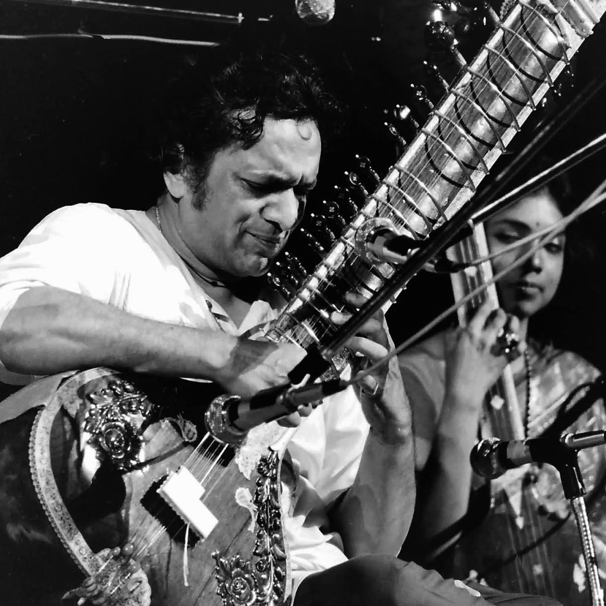 A black-and-white concert photo of a seated musician playing a sitar in the foreground while another musician plays a string instrument behind him.