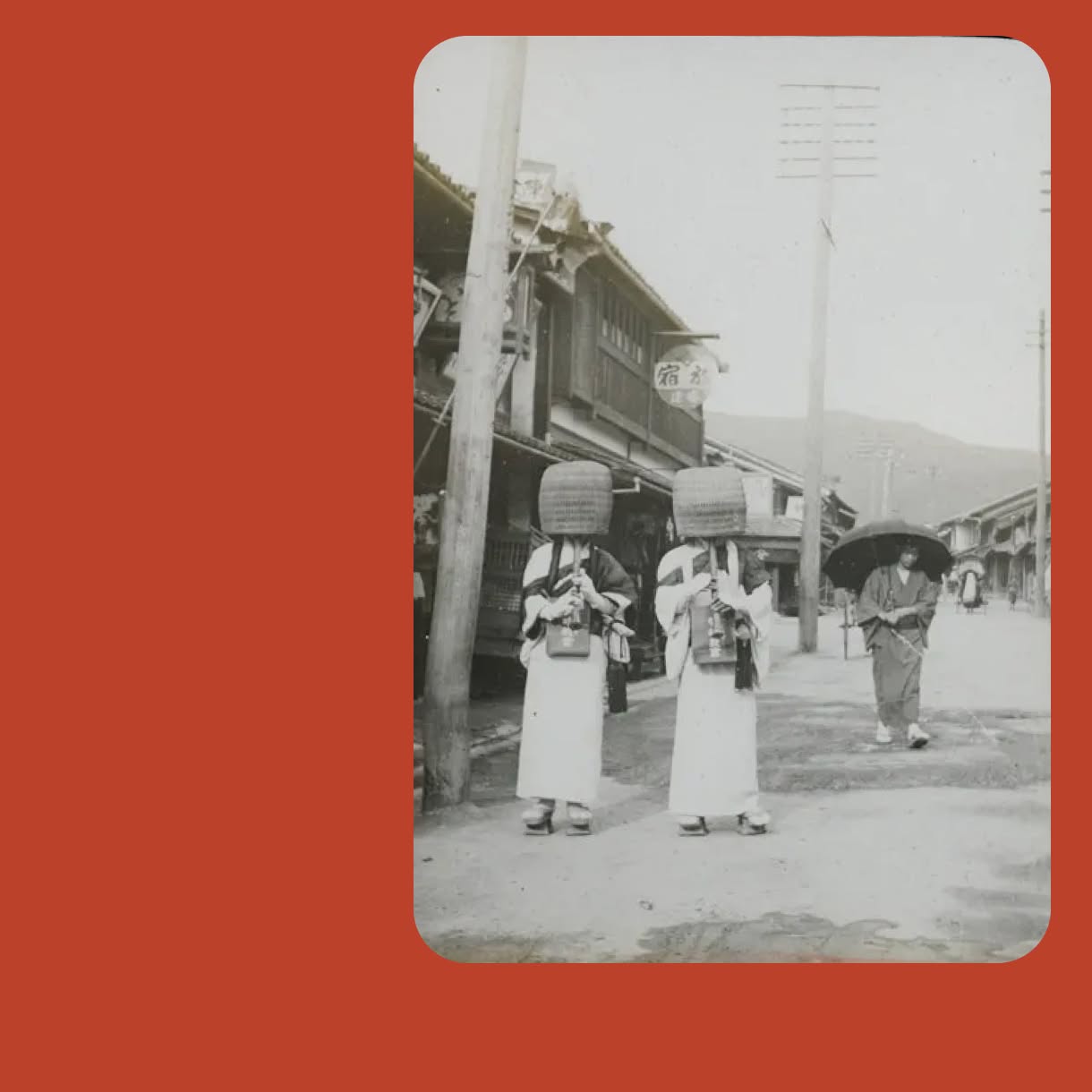 A black-and-white historical street photograph shows two people in light robes and large woven basket hats standing before wooden buildings, with another person carrying an umbrella behind them. The photo is framed by a solid blue background.
