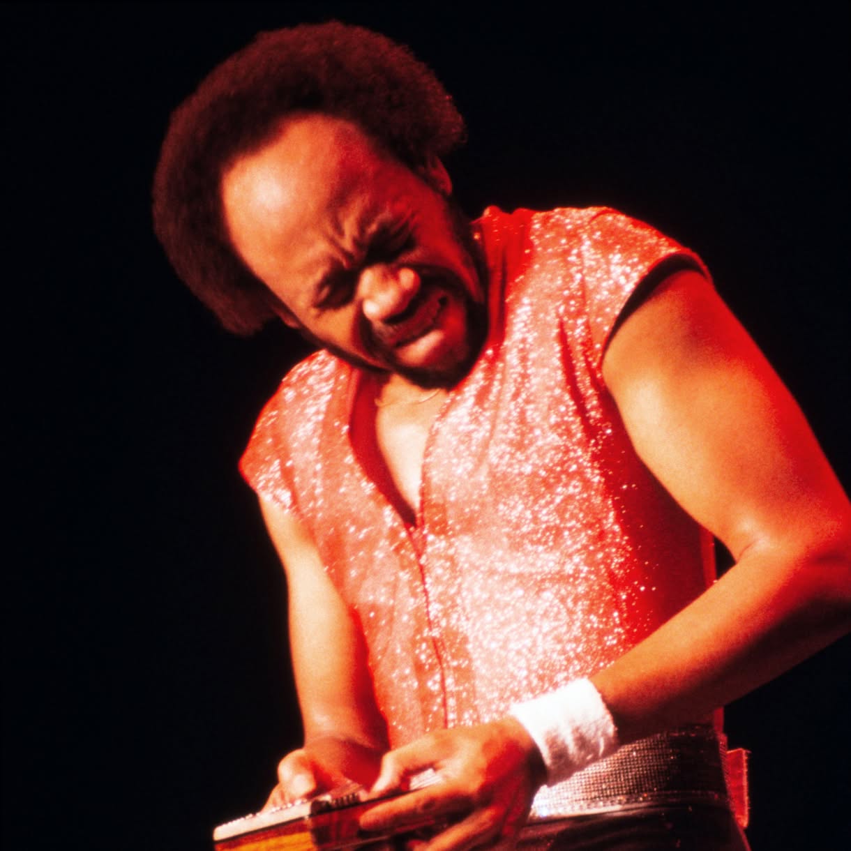 A male musician in a glittering red sleeveless top performs under red stage lighting, leaning over a small stringed instrument held at his waist.