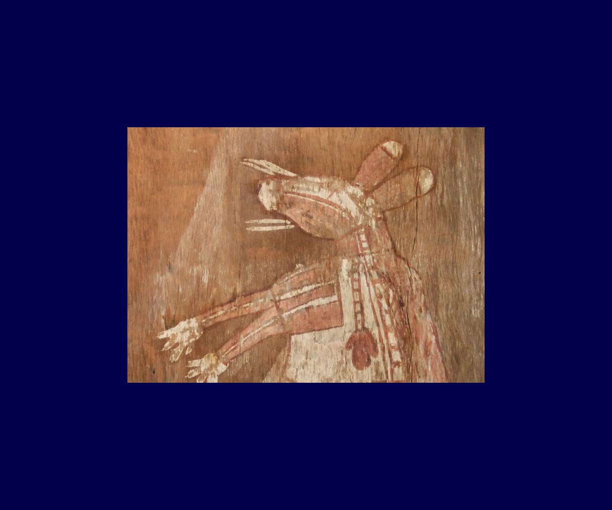 A faded reddish-brown rock painting depicts a stylized human-like figure with an elaborate headdress and outstretched arms, centered on a dark blue background.