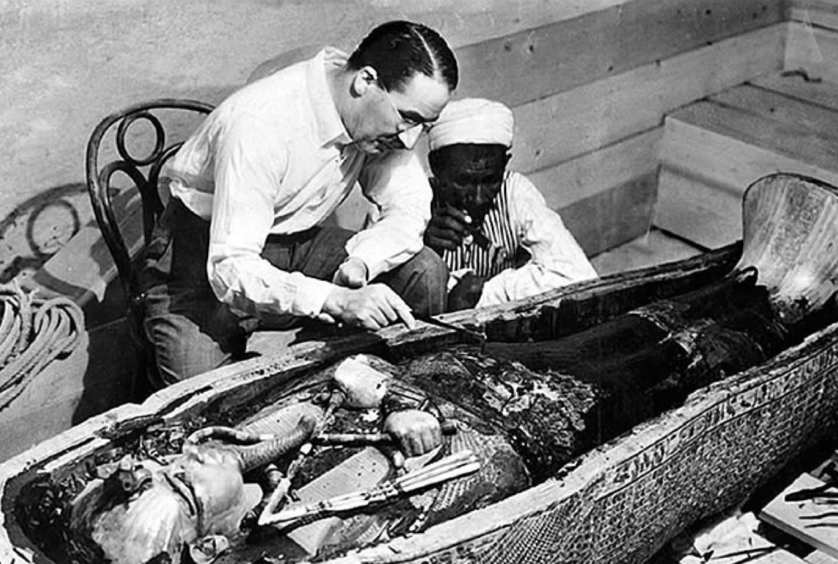 A black-and-white photograph shows two men examining an opened decorated sarcophagus containing a wrapped body and funerary objects.