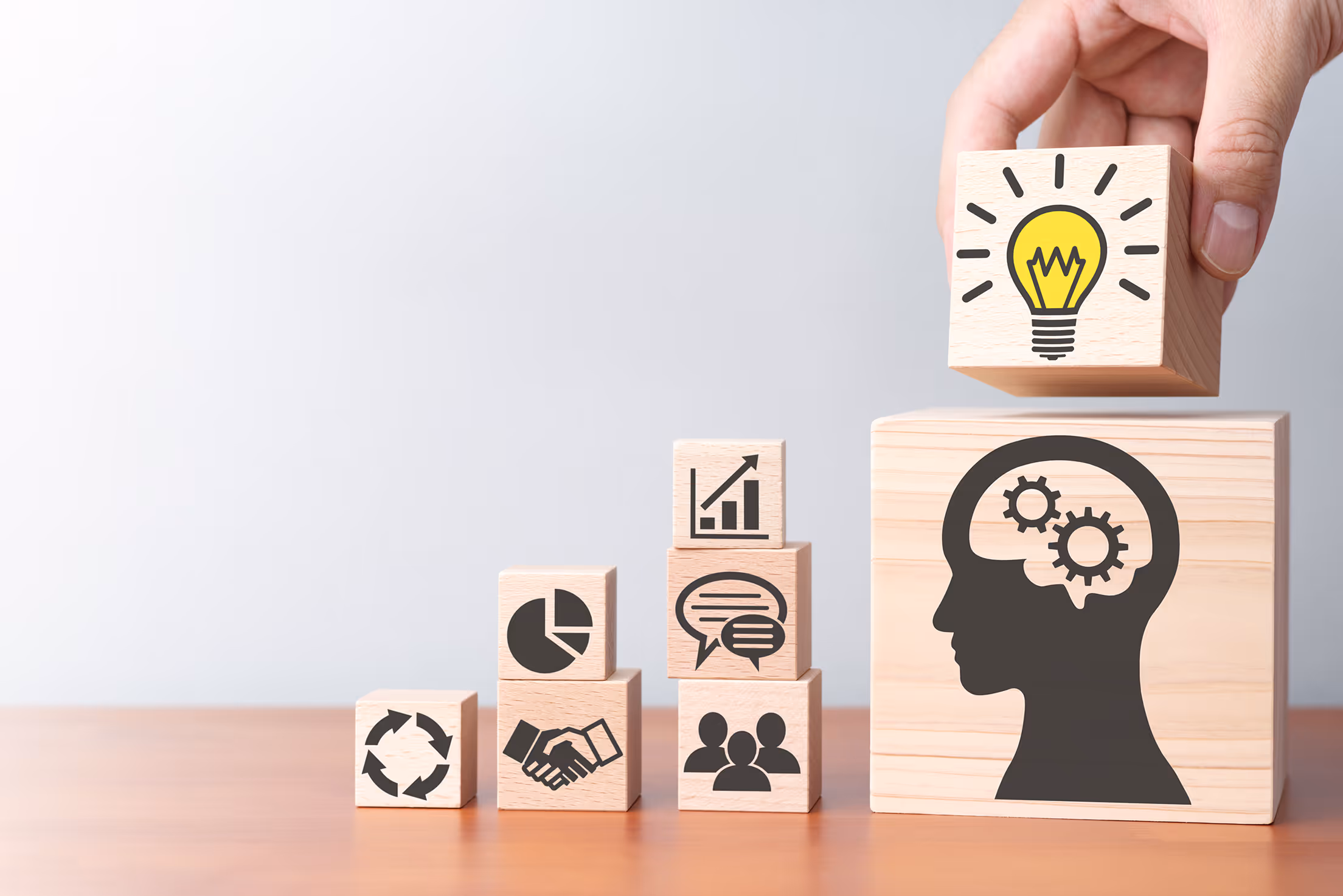Eine Hand legt einen Holzblock mit einem Glühbirnen-Symbol auf einen großen Block, der einen Kopf mit Zahnrädern zeigt, umgeben von aufsteigenden Blöcken mit Geschäfts- und Kommunikationssymbolen.
