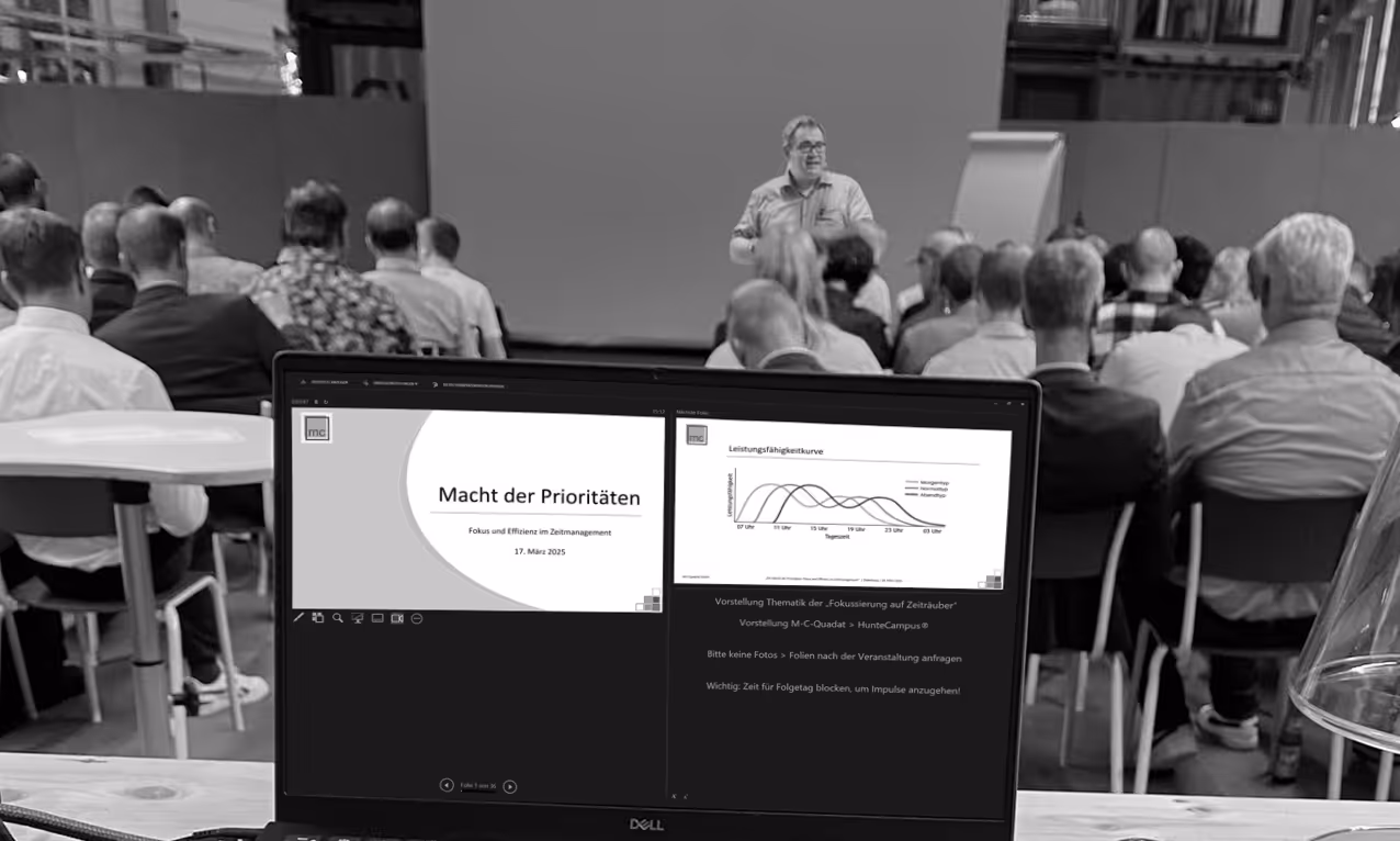 Zuhörer, die einem Referenten (Christian Liebezeit) bei einem Zeitmanagement-Seminar mit dem Titel „Macht der Prioritäten“ zusehen, während auf dem Laptop-Bildschirm Folien gezeigt werden.