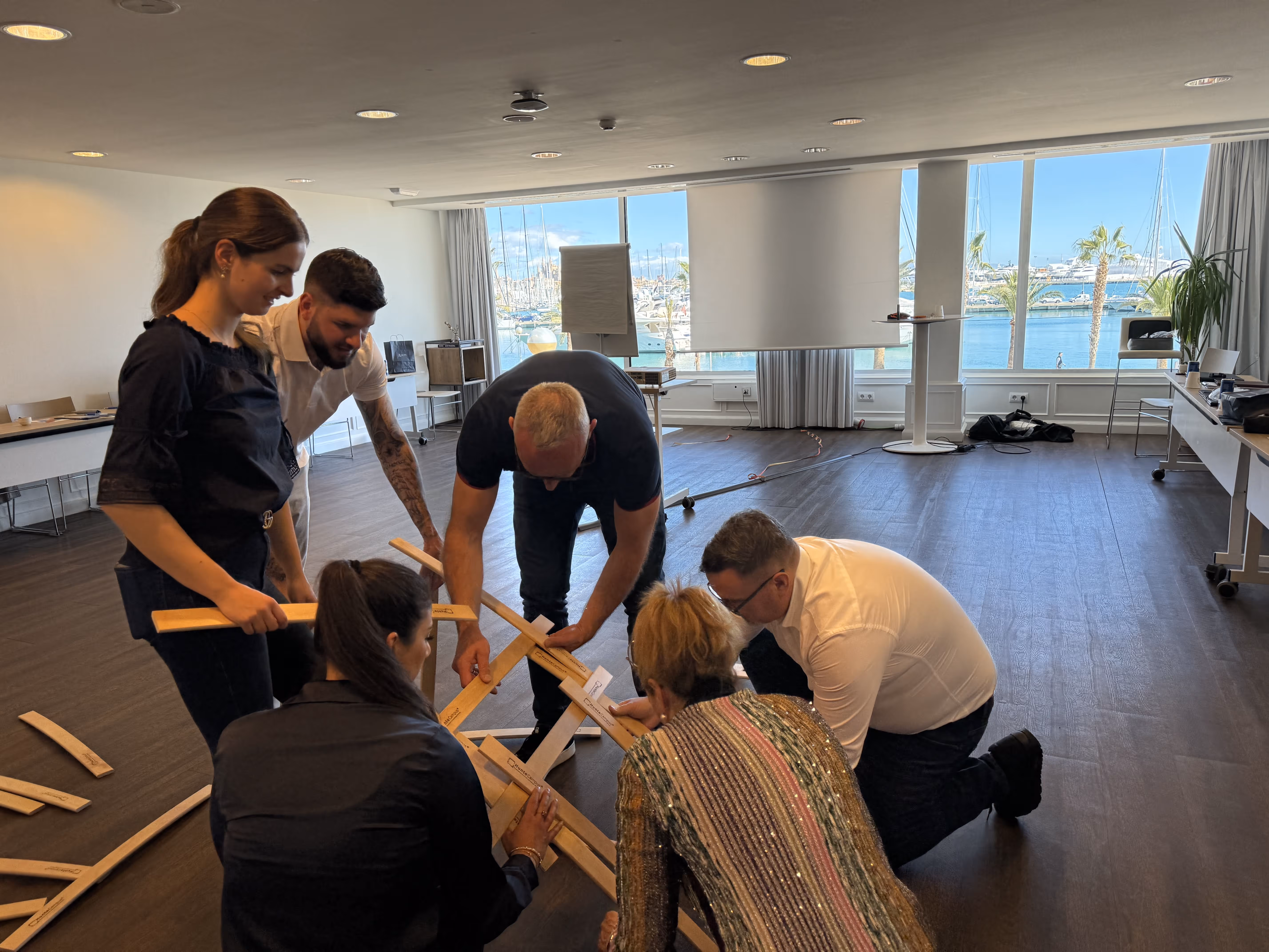 Sechs Erwachsene, die gemeinsam Holzstäbe zusammenbauen, in einem hellen Raum mit großen Fenstern, von denen aus man auf einen Yachthafen mit Booten und Palmen blickt.