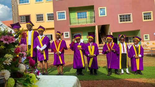 A group of nine children wearing purple and yellow graduation gowns and caps, standing outside on a green patch with a building in the background.