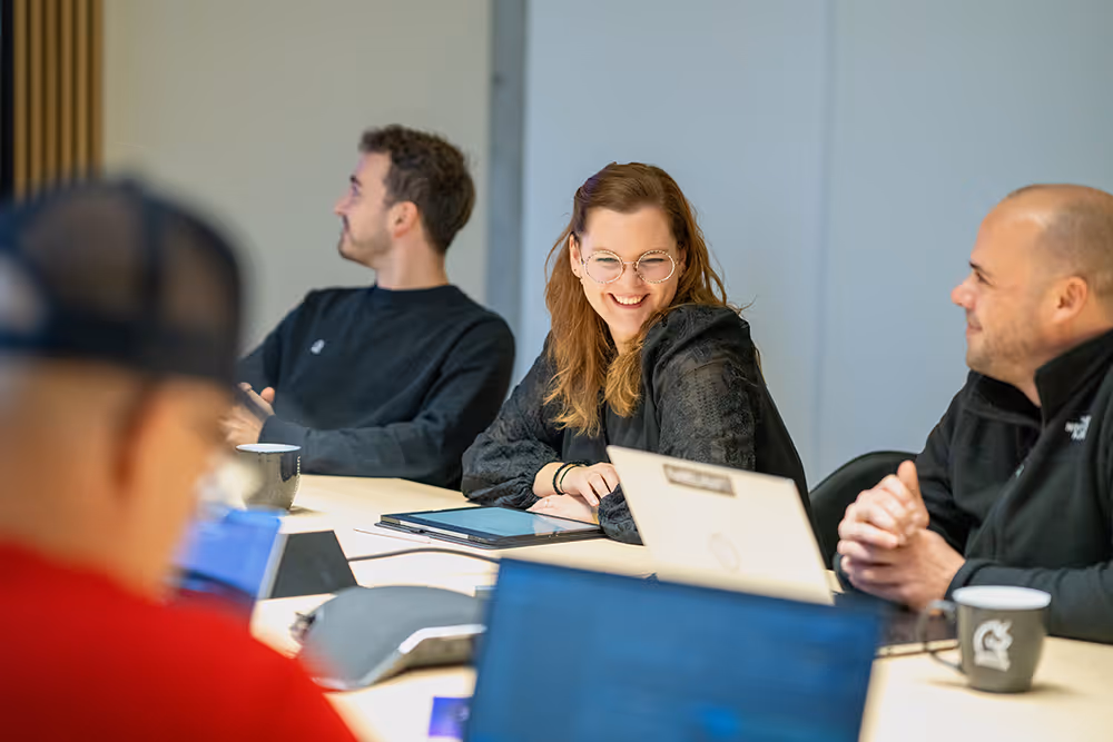 Drei Personen sitzen an einem Konferenztisch mit Laptops und Tassen, während eine Frau in der Mitte lächelt.