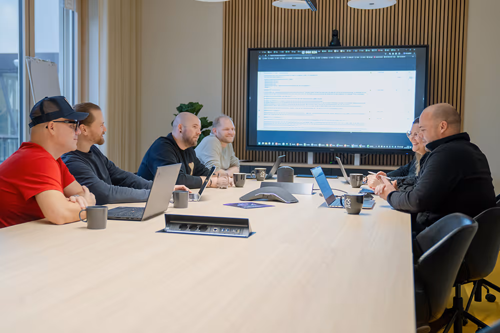 Sechs Personen sitzen an einem langen Konferenztisch, mit geöffneten Laptops und Kaffeetassen, während sie auf einen großen Bildschirm schauen.