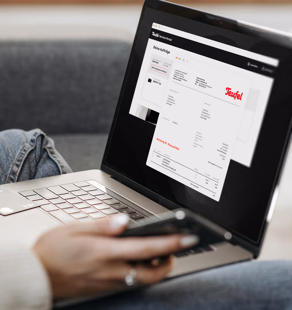 Hand hält Smartphone vor einem Laptop, auf dessen Bildschirm das Teufel Service-Portal mit einer Rechnung angezeigt wird.