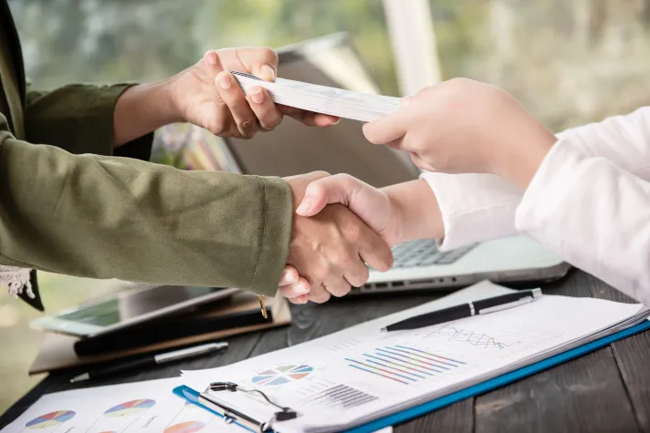Cette image montre deux personnes échangeant une poignée de main ainsi qu'une liasse de documents 