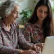 Cette image montre une femme âgée accompagnée d'une autre plus jeune, devant un écran d'ordinateur