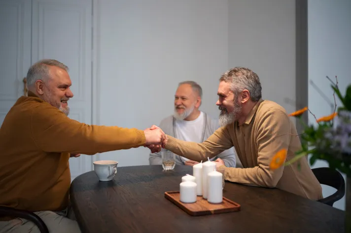 Cette image montre trois hommes seniors autour d'une table, souriant et échangeant une poignée de main