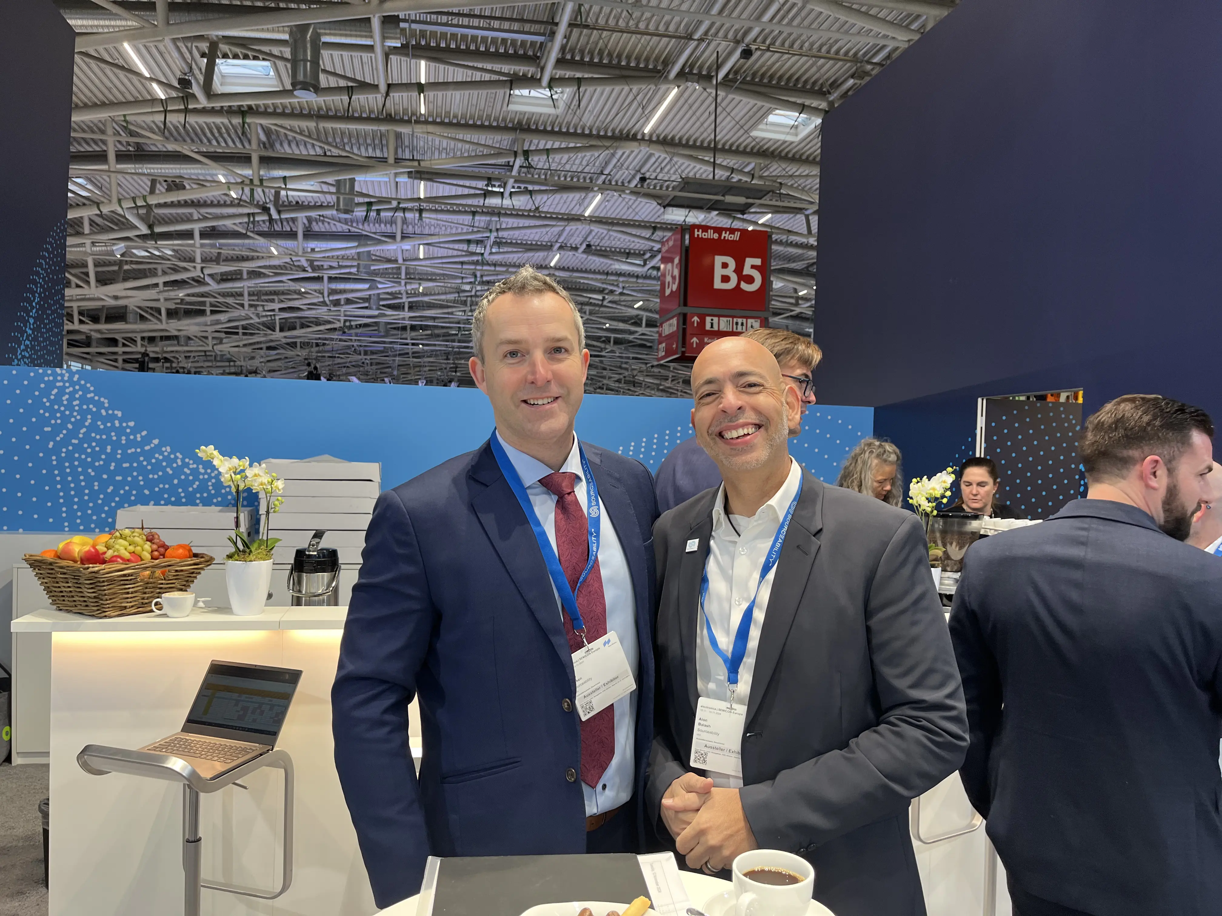 Two men in suits smiling at a trade show booth with coffee and fruit in the background.