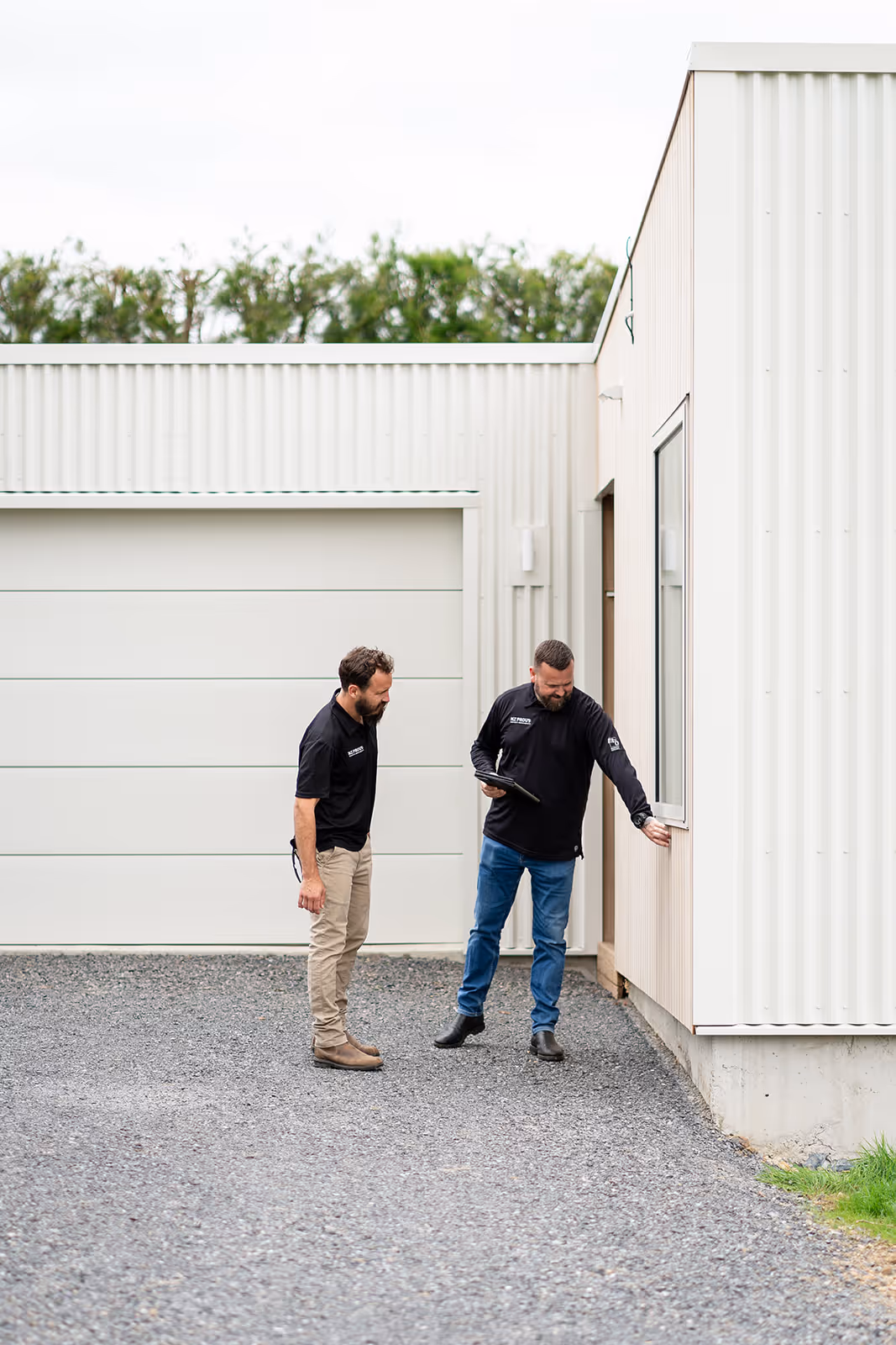 Two men in black shirts inspecting the exterior wall of a modern white corrugated metal building with a gravel driveway.