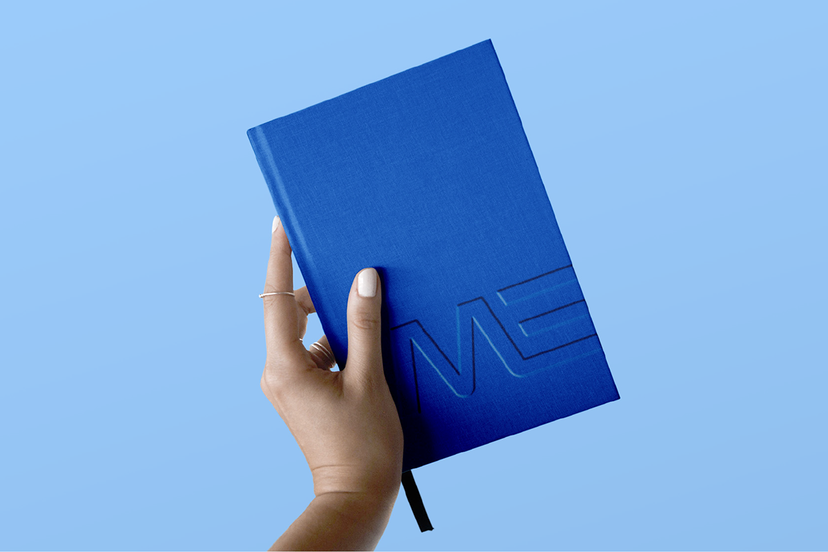 A hand with white nail polish and rings holding a closed blue notebook embossed with the letters 'ME' on a light blue background.
