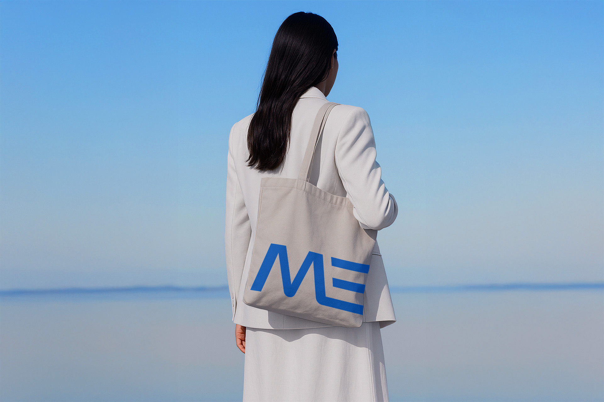 Woman dressed in a white blazer and skirt standing with her back to the camera, holding a beige tote bag with a blue stylized logo against a calm blue sky and water background.