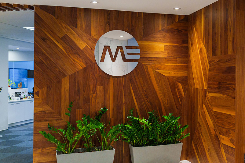 Indoor office area with a wood-paneled wall featuring a circular logo with the letters M and E, and two white rectangular planters filled with green leafy plants in front.