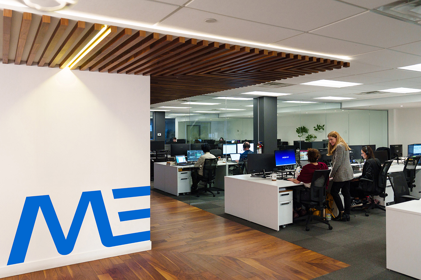 Modern office space with wooden floor, white desks, and glass partitions; several people working at computers; one woman standing and talking to a seated colleague.