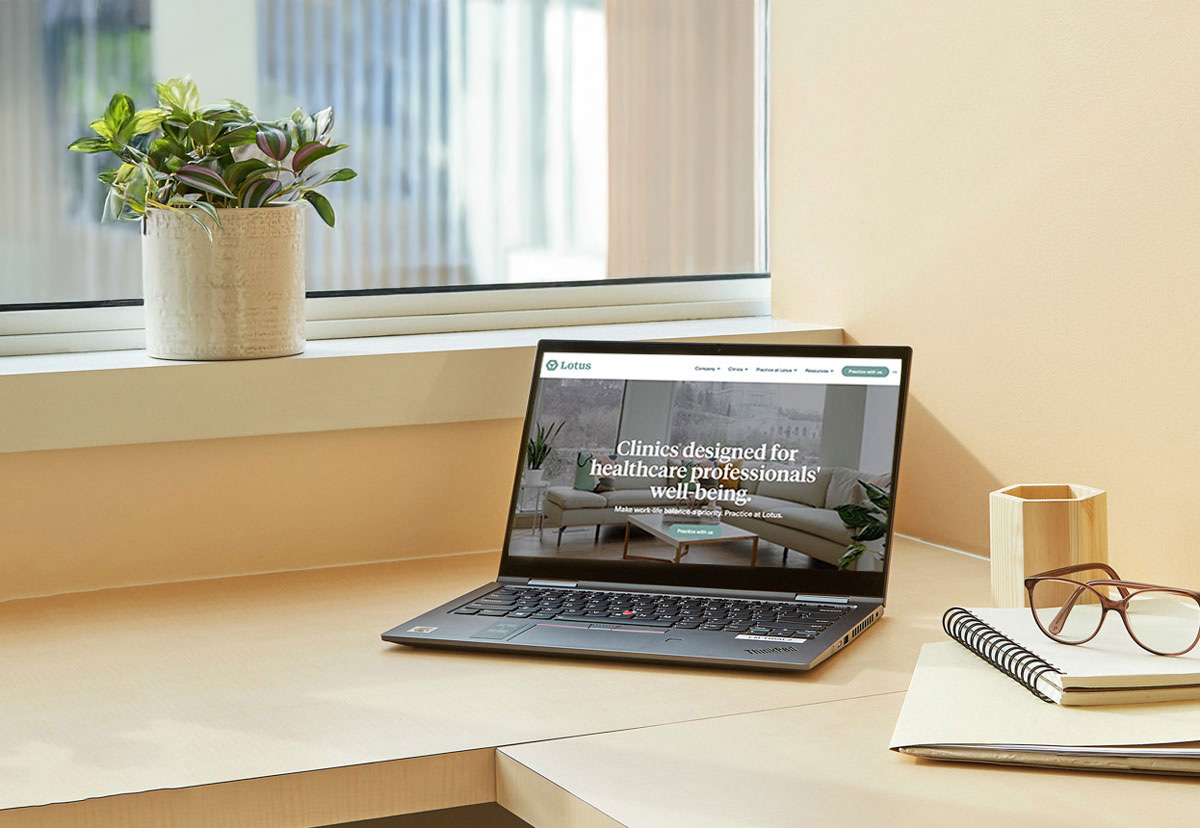 A clean, light wood desk with a laptop showing a healthcare clinic website, a potted plant on the windowsill, a pair of glasses, a wooden pen holder, and notebooks.