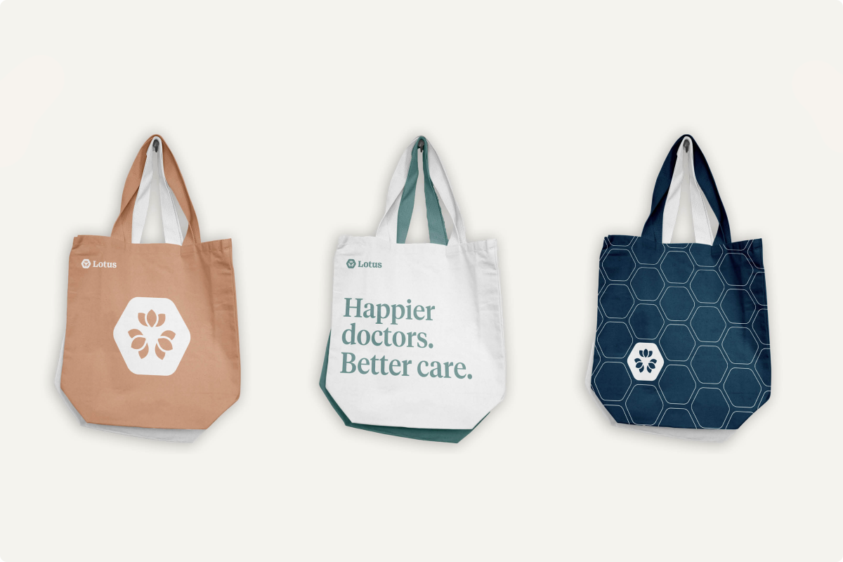 Three tote bags on a light background: left bag is light brown with white lotus flower logo and 'Lotus' text; middle bag is white with green handles and text 'Happier doctors. Better care.' and a small green Lotus logo; right bag is dark blue with a hexagonal pattern and a small white lotus logo inside one hexagon, with dark handles.