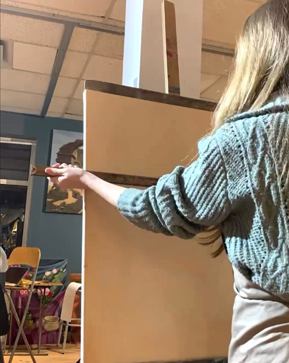 Young woman with long blonde hair wearing a knitted teal sweater and apron holding a paintbrush and facing a blank canvas and easel in an indoor studio setting.