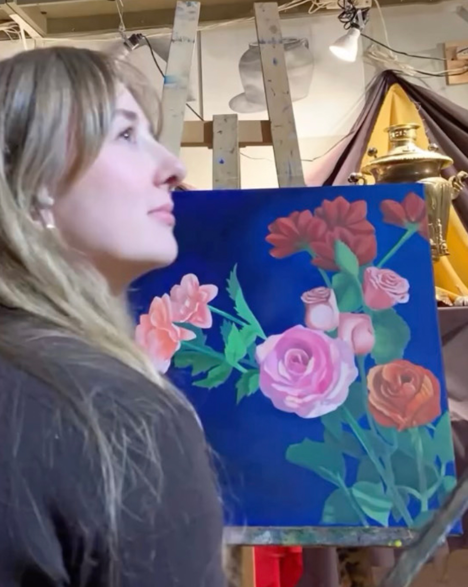 Woman with long blonde hair looking at a colourful floral painting of roses and other flowers on a blue background, set on an easel in an indoor art studio with draped fabrics and a brass samovar in the background.
