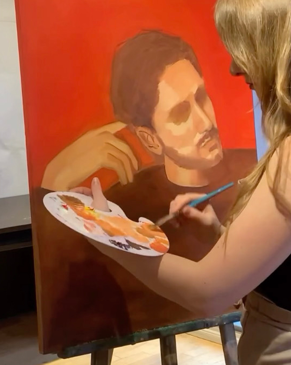 Artist painting a portrait of a man with brown hair and a beard on a canvas with a red background, holding a palette of paint and a brush.