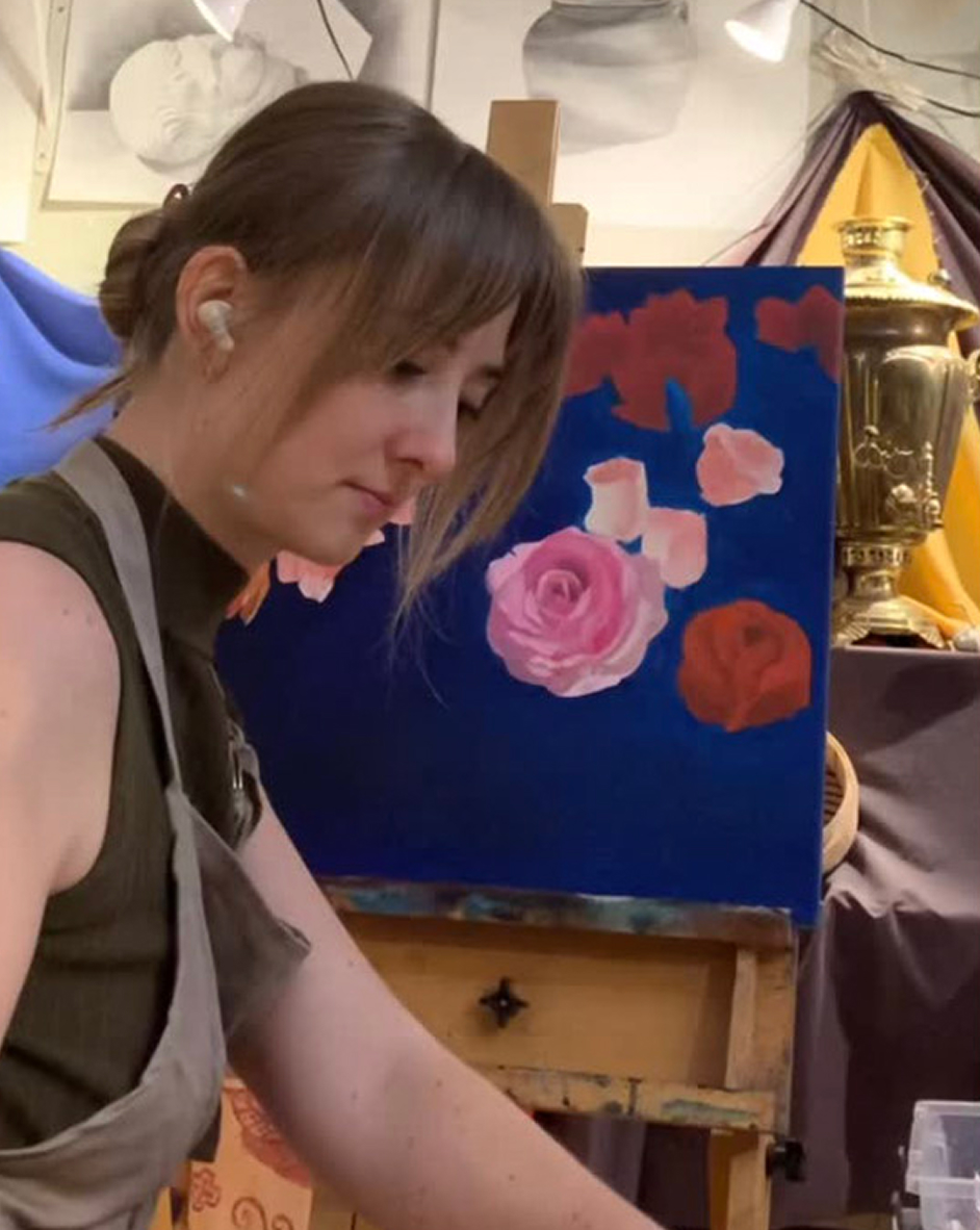 A woman with blonde hair tied back wearing a sleeveless top and apron is focused on painting flowers on a canvas with a dark blue background in an art studio.