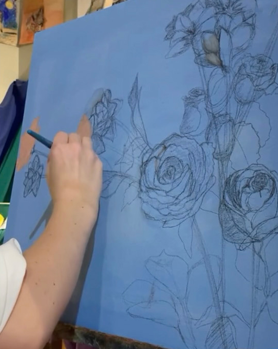 Artist's hand holding a pencil while sketching detailed flowers and leaves on a blue canvas.