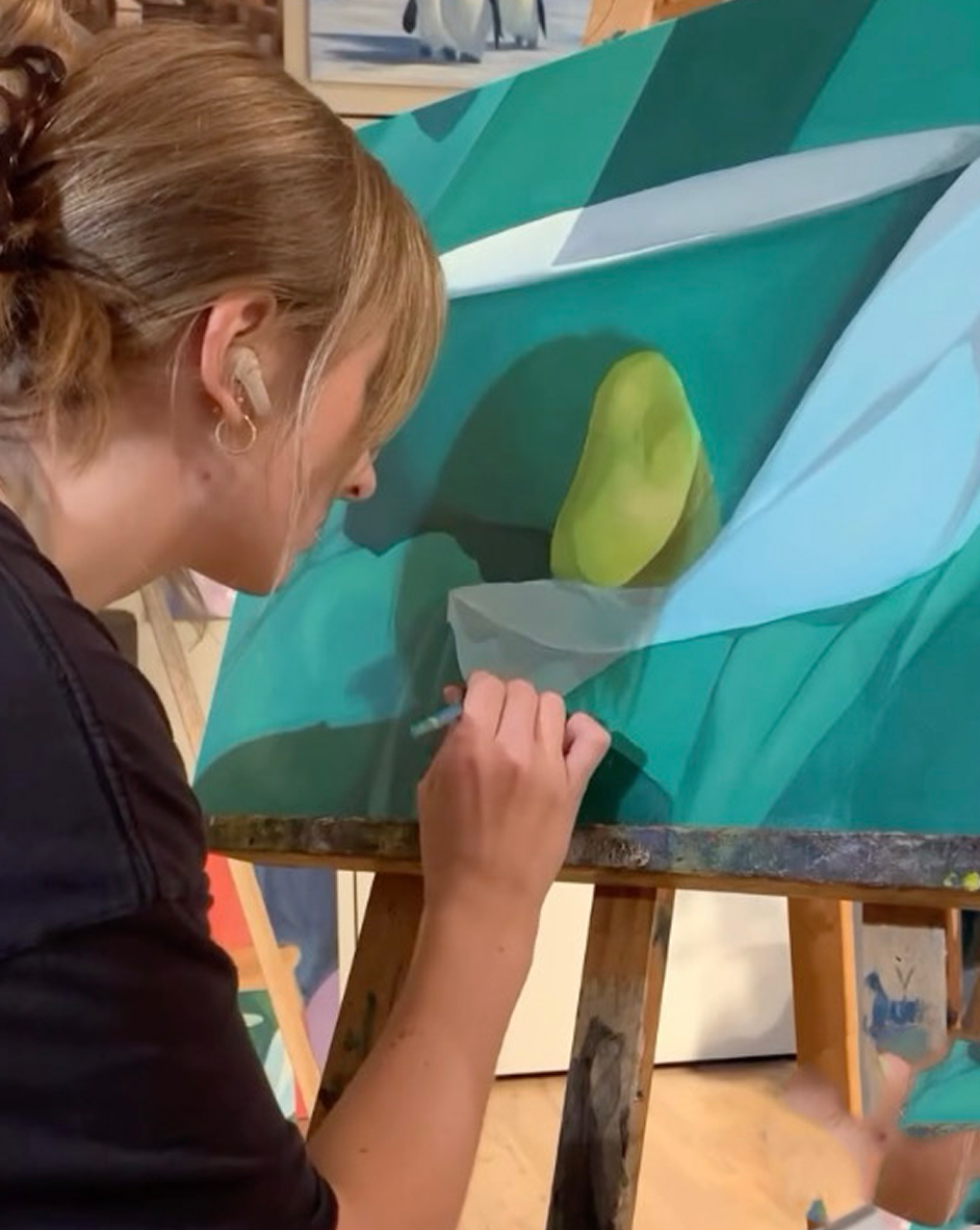 A person with blonde hair tied back, wearing a black shirt, painting an artwork with green and blue shades on a canvas set on an easel indoors.