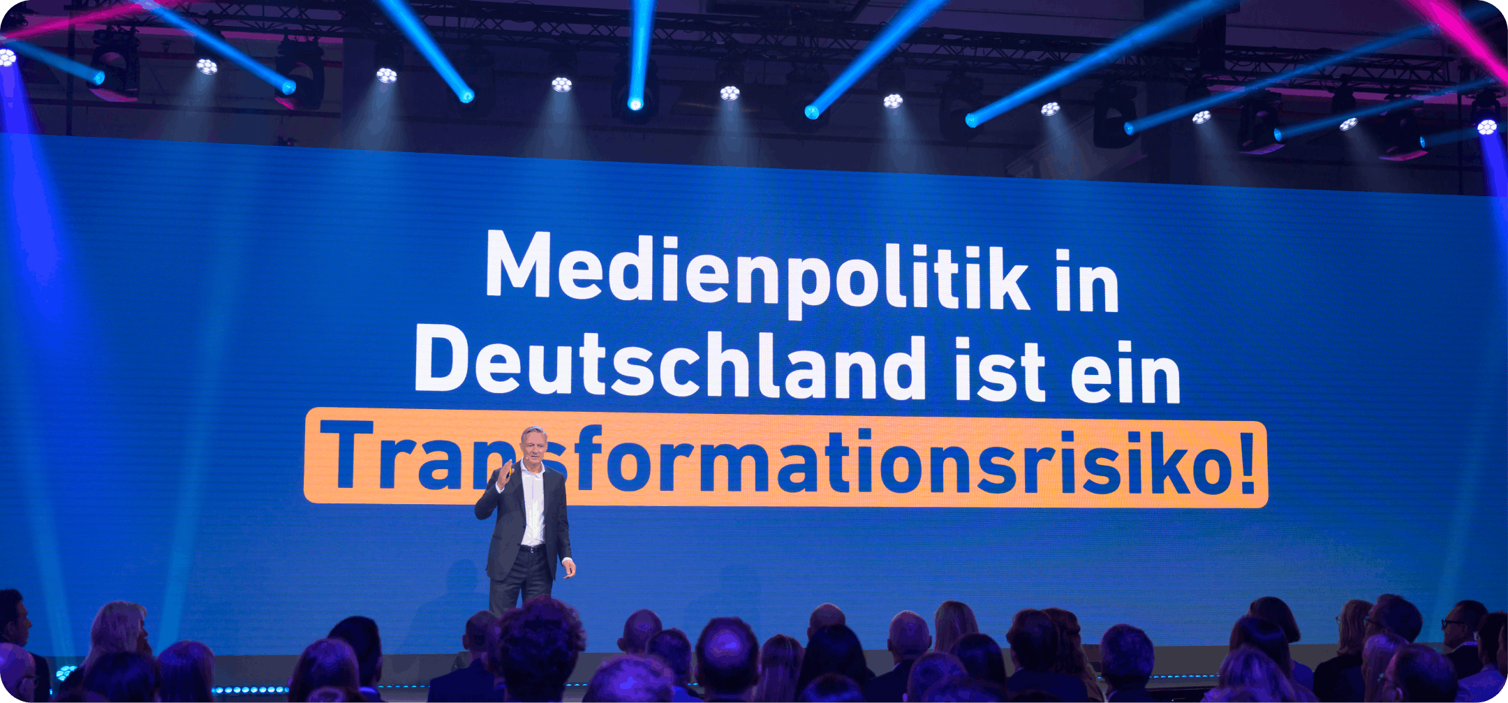Ein Redner steht auf der Bühne vor dem Publikum, hinter ihm ist auf einer großen Leinwand der Text „Medienpolitik in Deutschland ist ein Transformationsrisiko!“ zu sehen.