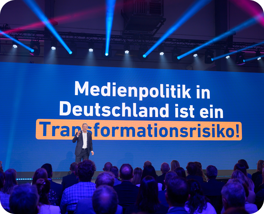 Ein Redner steht auf der Bühne vor dem Publikum, hinter ihm ist auf einer großen Leinwand der Text „Medienpolitik in Deutschland ist ein Transformationsrisiko!“ zu sehen.