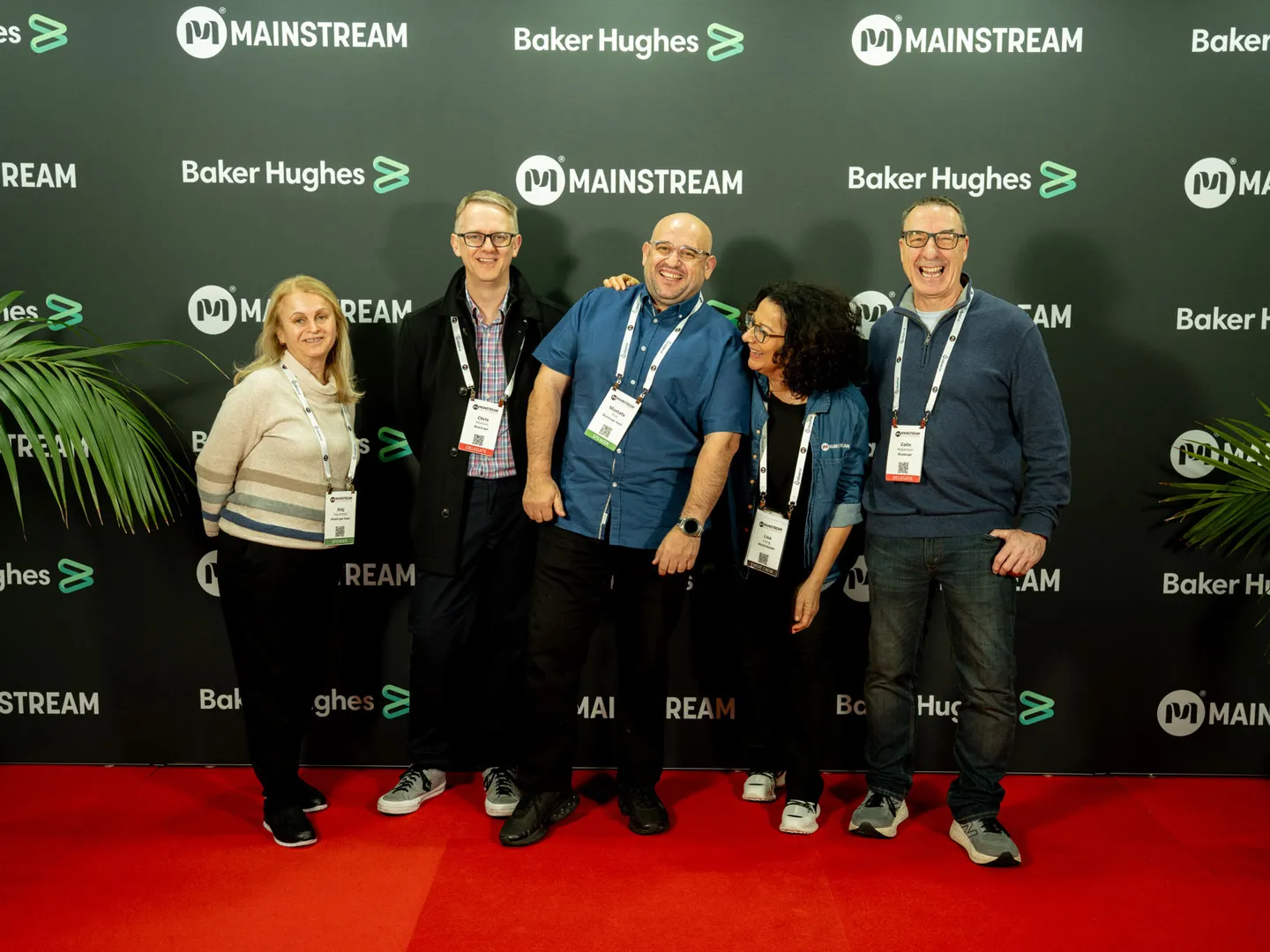 Five people wearing conference badges standing together smiling on a red carpet in front of a backdrop with Mainstream and Baker Hughes logos.