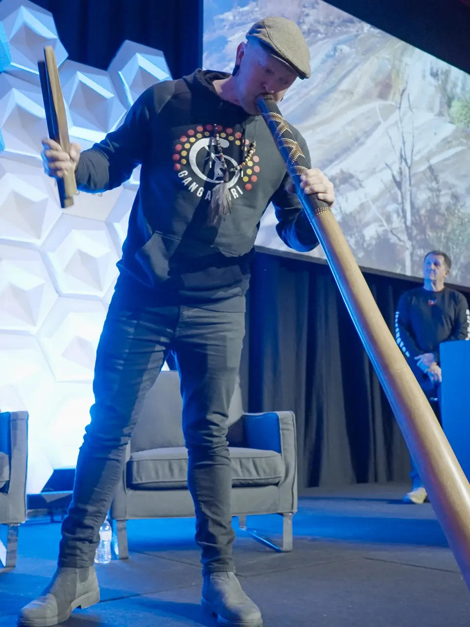 Man playing a didgeridoo on a stage, holding a wooden percussion instrument in one hand