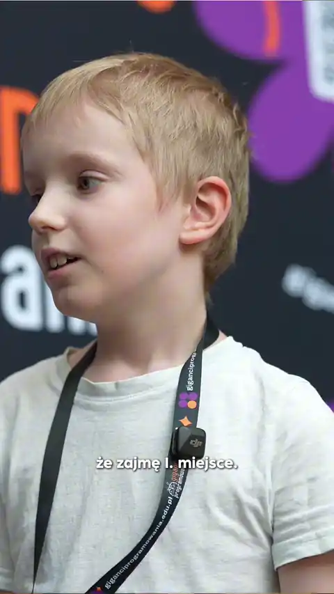 Young boy with short blonde hair wearing a white shirt and a black lanyard with a microphone clip, looking to the side.