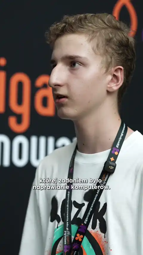 Young man with light curly hair wearing a white shirt and a lanyard, standing in front of a blurred background with orange and white text.