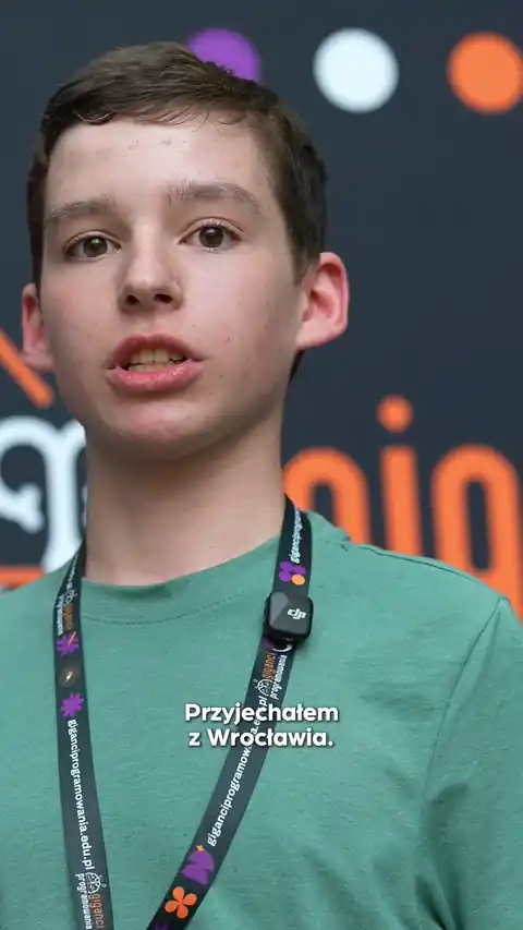 Young person with short brown hair wearing a green shirt and a black lanyard with colorful icons, speaking in front of a blurred background with orange and white text.