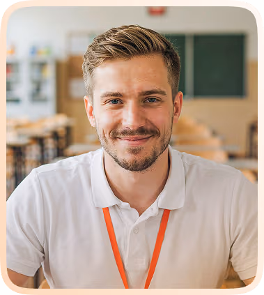 Smiling young man with short light brown hair, beard, and mustache wearing a white polo shirt and an orange lanyard sitting in a bright, modern classroom with blurred desks and a chalkboard in the background.