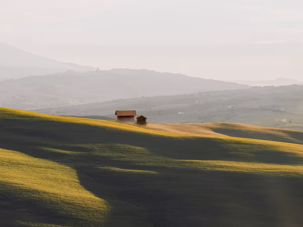 Two small wooden cabins on gently rolling green hills with a hazy mountain range in the background.