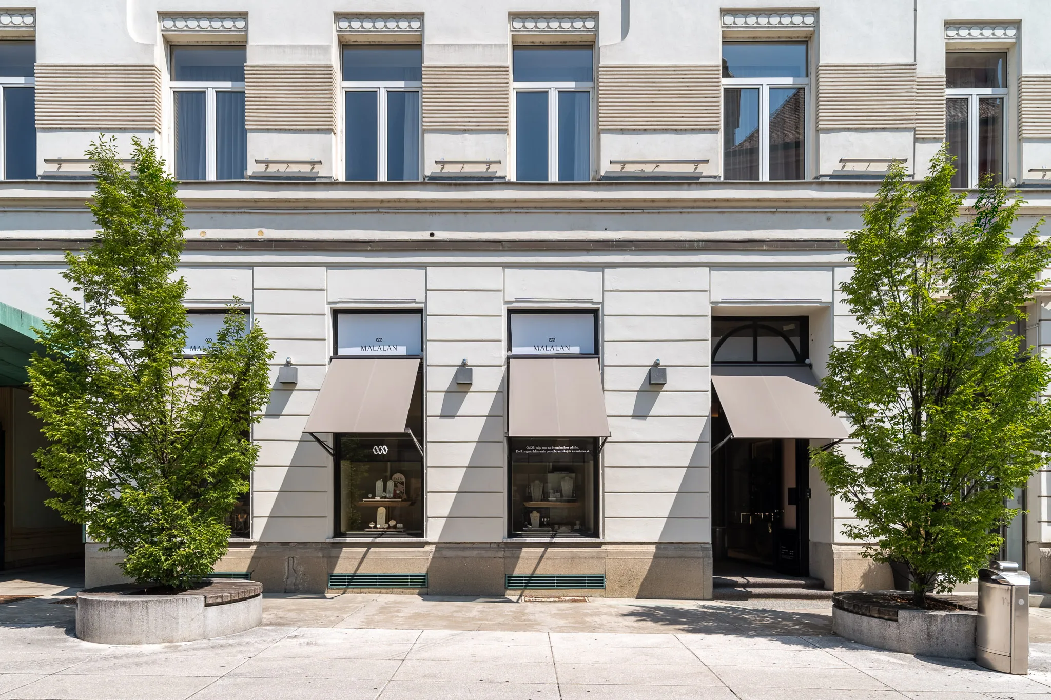 Street view of Malalan jewelry boutique in Ljubljana, with a classic stone façade, branded window displays, and taupe awnings, showcasing high-end retail design by Studio Mosh.