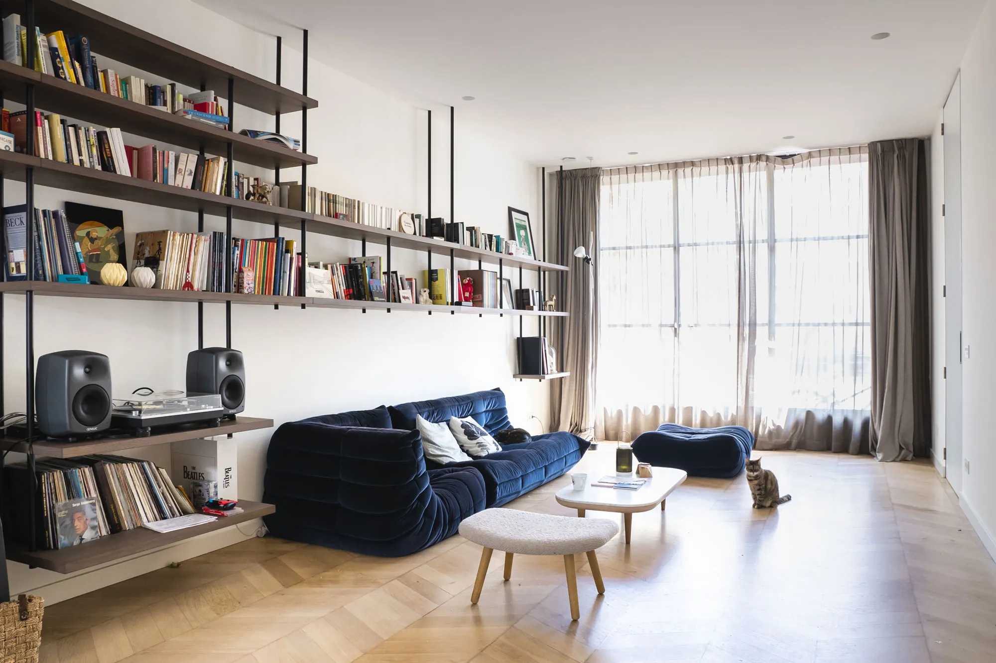 Modern living room interior with custom wall-mounted shelving and integrated bookcase design by Studio Mosh, featuring a deep blue velvet sofa, mid-century coffee tables, and natural light filtering through sheer curtains — minimalist yet eclectic apartment design.