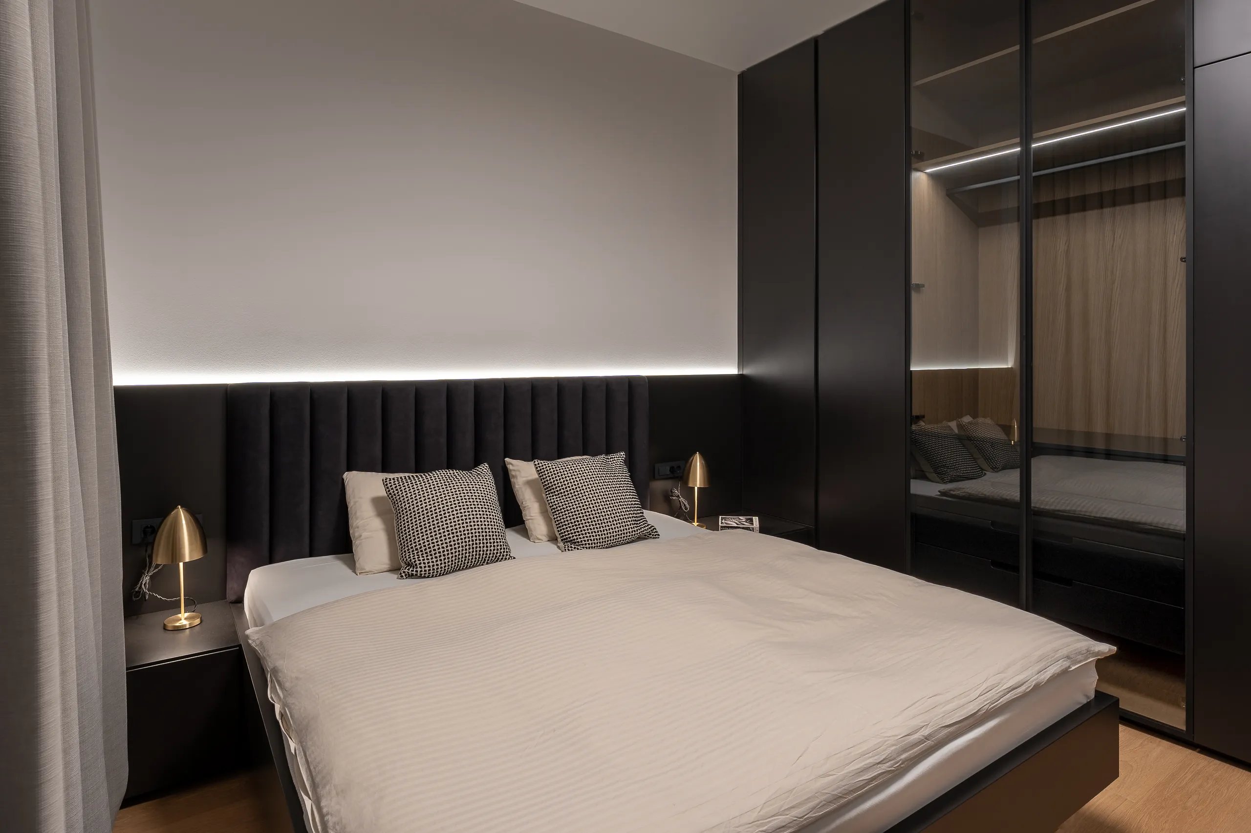 Minimalist bedroom with black panel headboard, soft neutral bedding, golden bedside lamps, and black glass wardrobe – modern sleeping space with warm lighting.