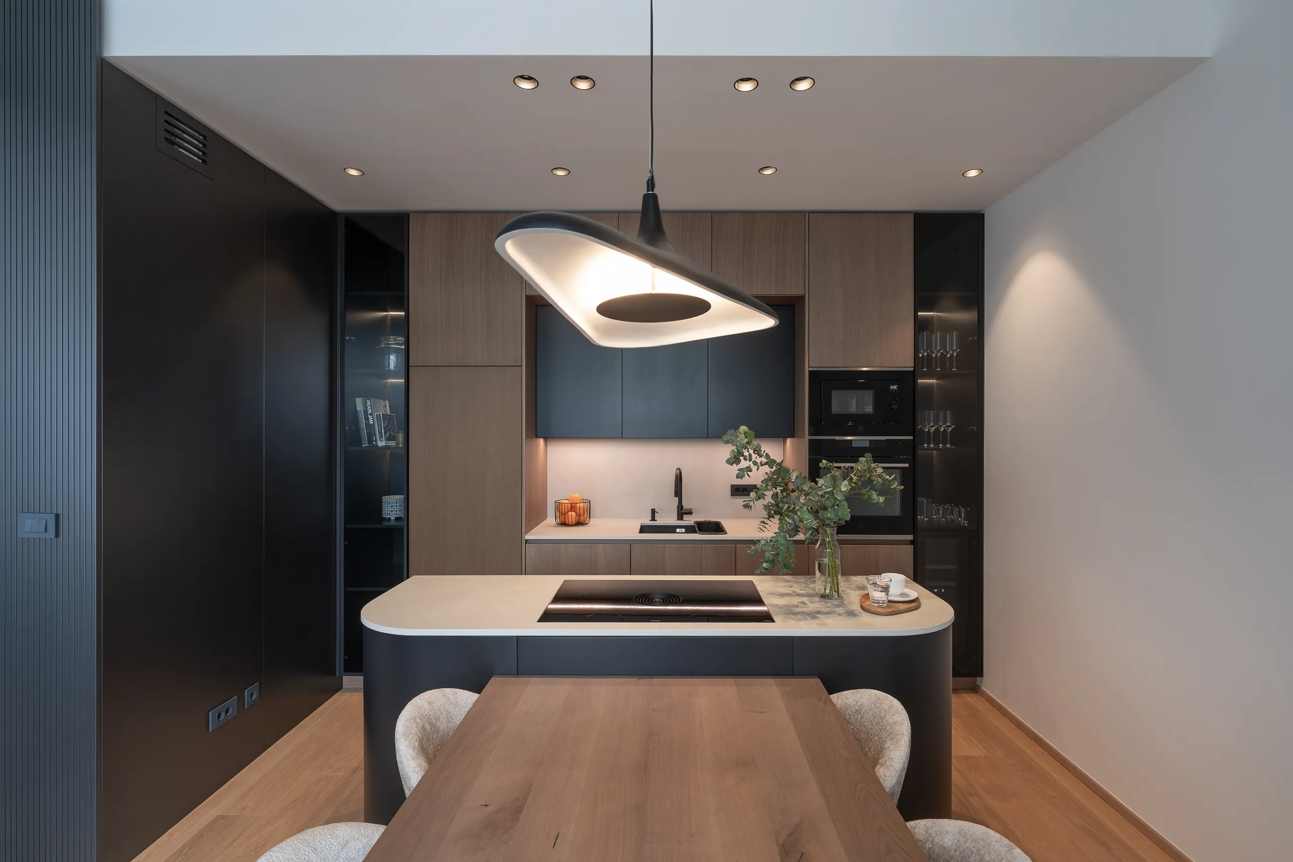 Frontal view of luxury kitchen with integrated appliances, black and oak cabinetry, curved island, and designer pendant light – high-end apartment kitchen.
