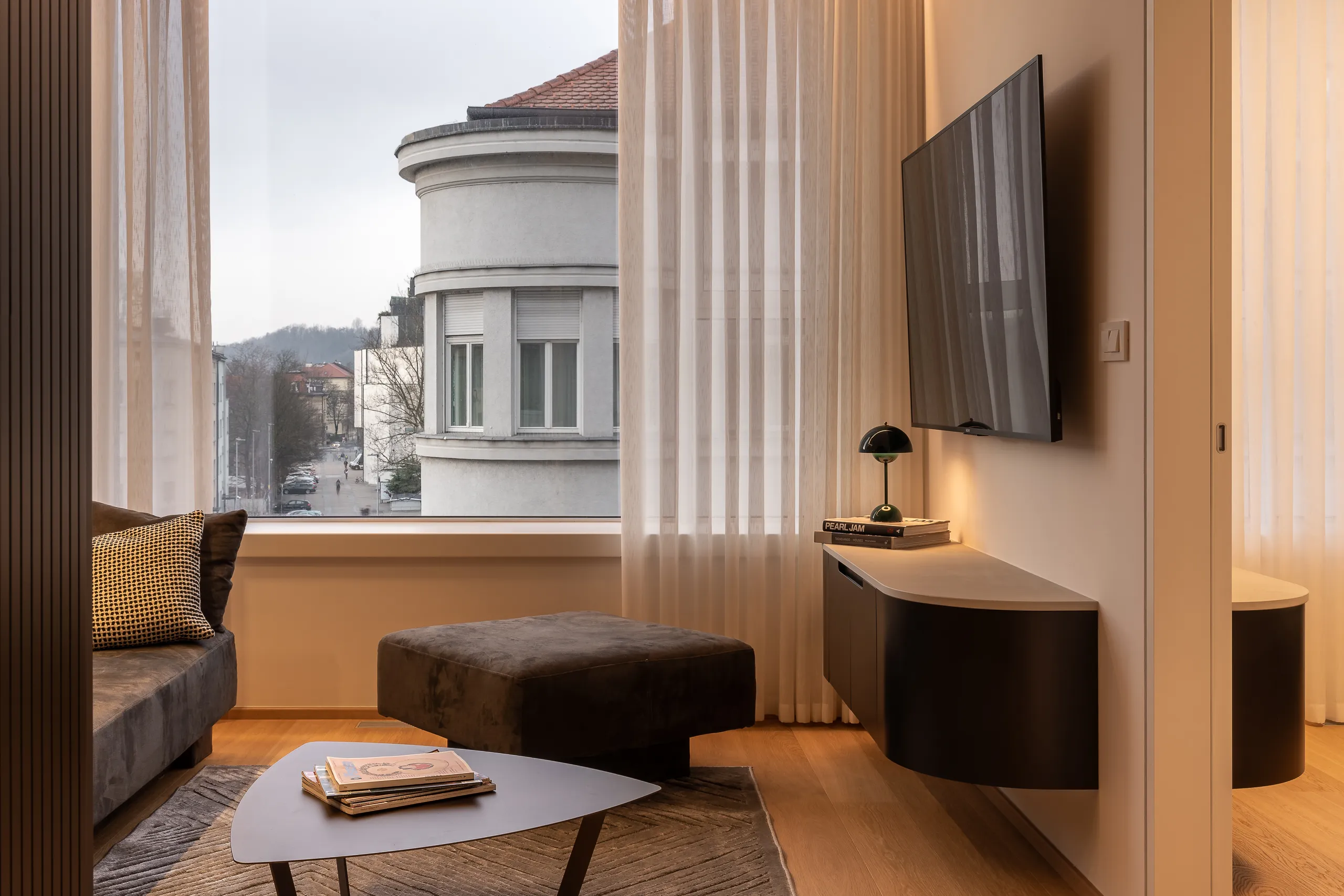 Cozy modern living room with wall-mounted TV, curved black console, ottoman, and large window with sheer curtains overlooking a European street.