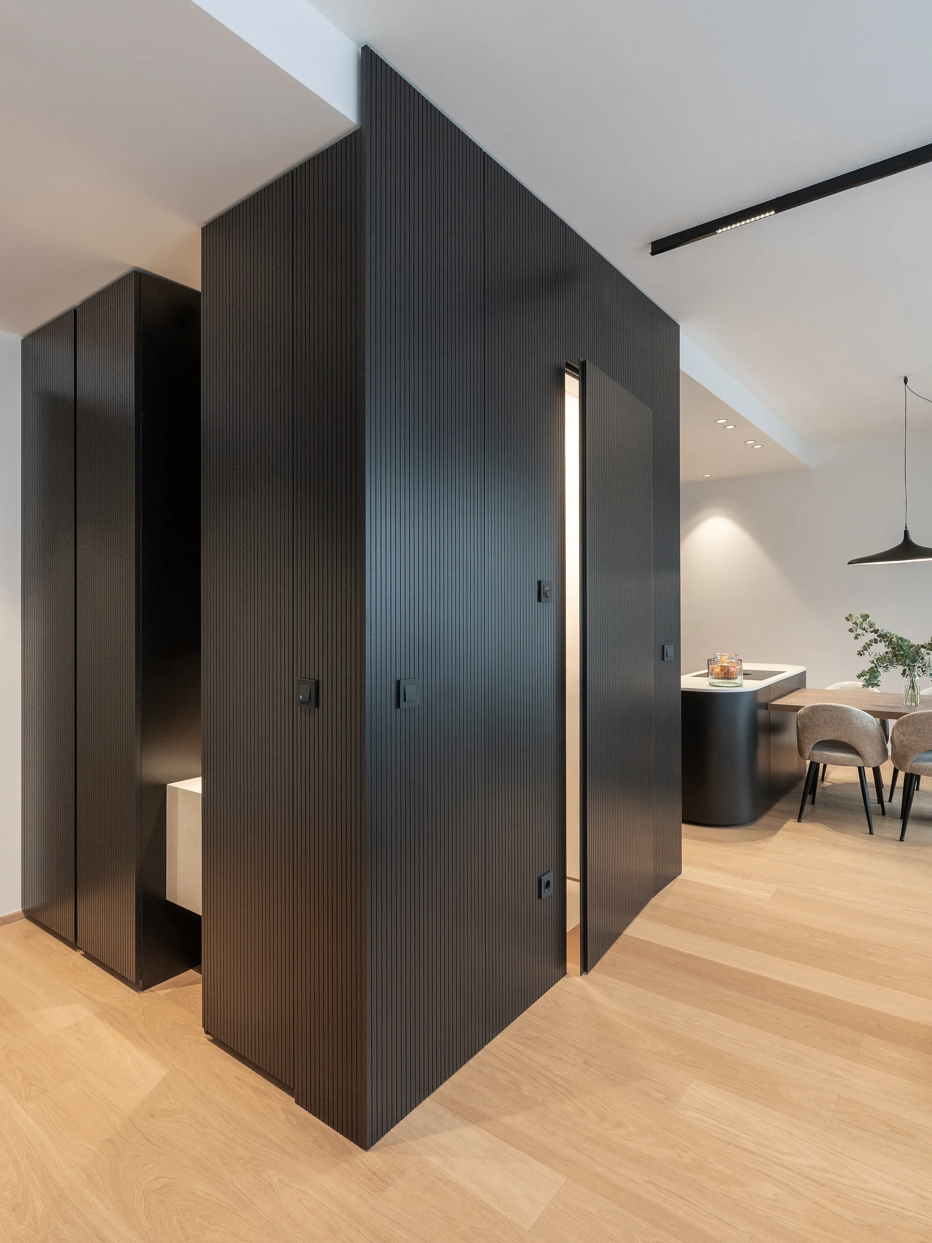 Open-plan view of a modern apartment showcasing black ribbed wood partitions, warm wood flooring, and stylish dining area connected to the kitchen.