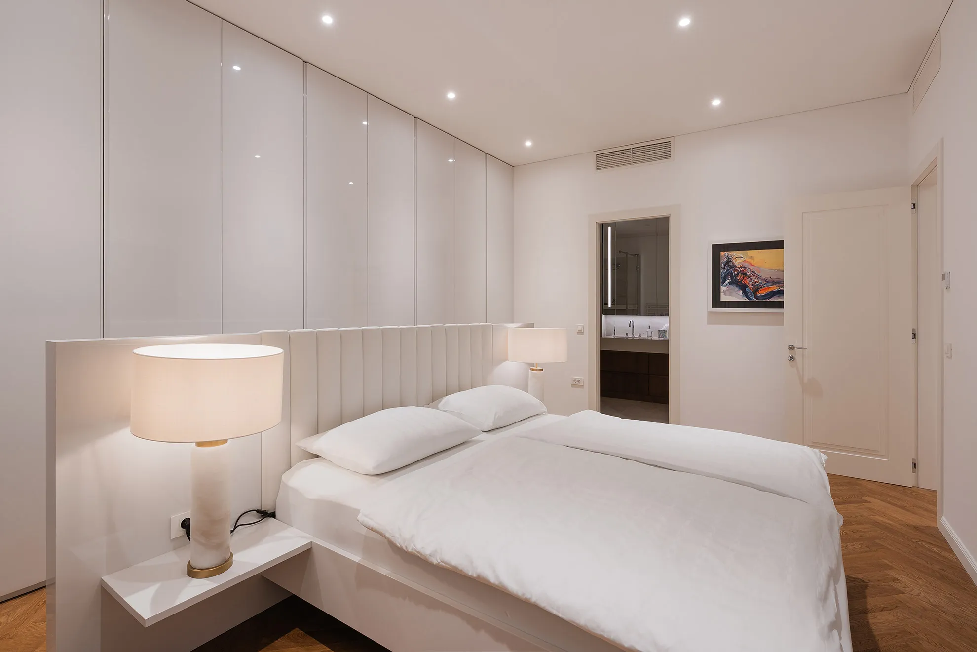 Minimalist and bright bedroom in Penthouse Schellenburg with sleek white furniture, ambient lighting, and an open layout that connects seamlessly to the en-suite bathroom.
