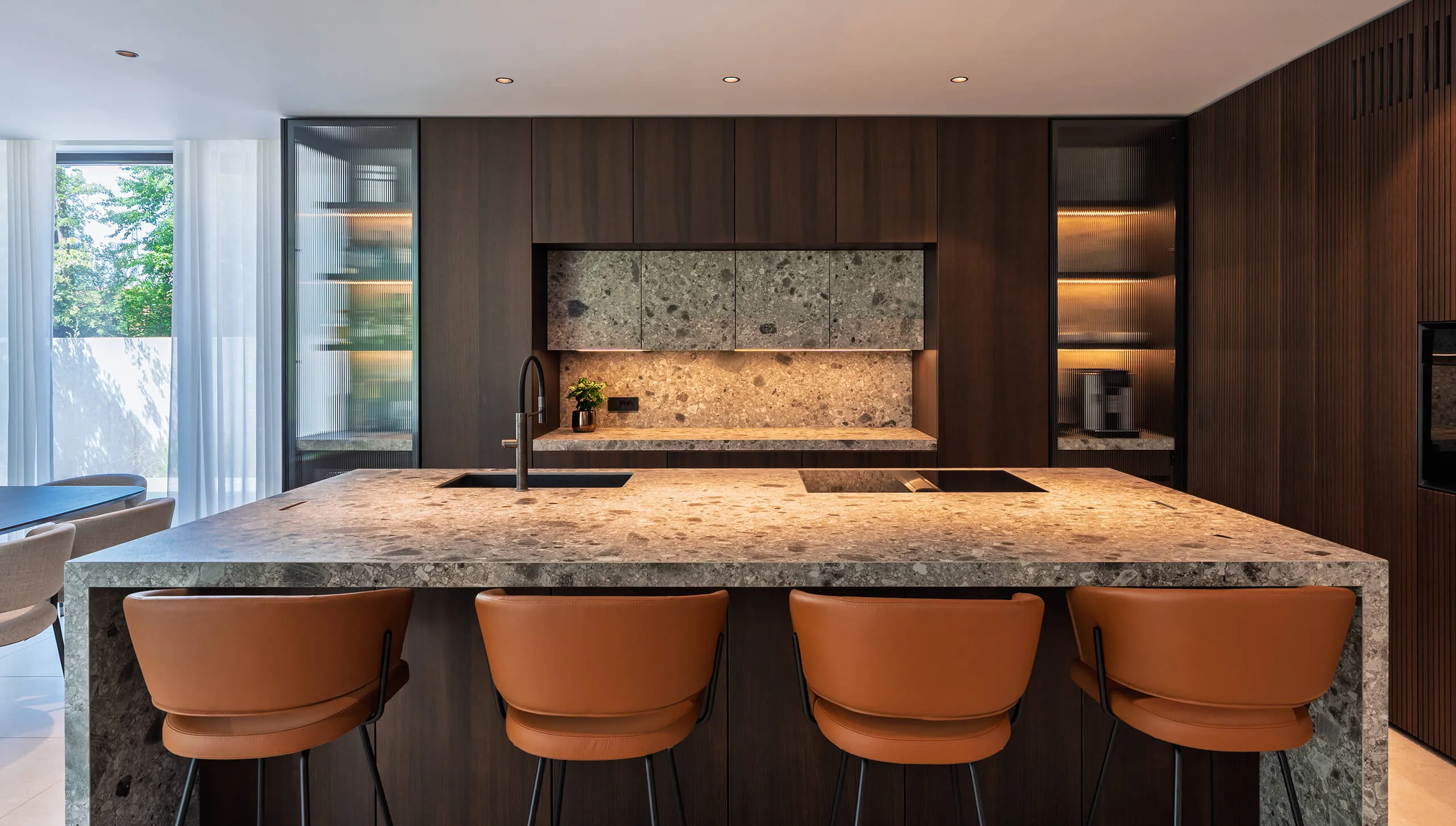 Open-plan living room and kitchen in a luxury mountain chalet by Studio Mosh, with exposed wooden beams, a marble island, rustic textures, and custom-built media wall — blending alpine comfort with modern interior design.
