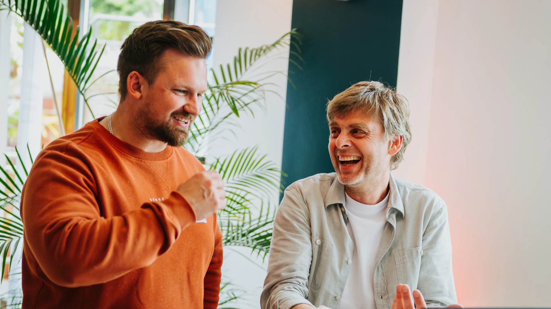 Two men smiling and talking in a bright room with green plants in the background.