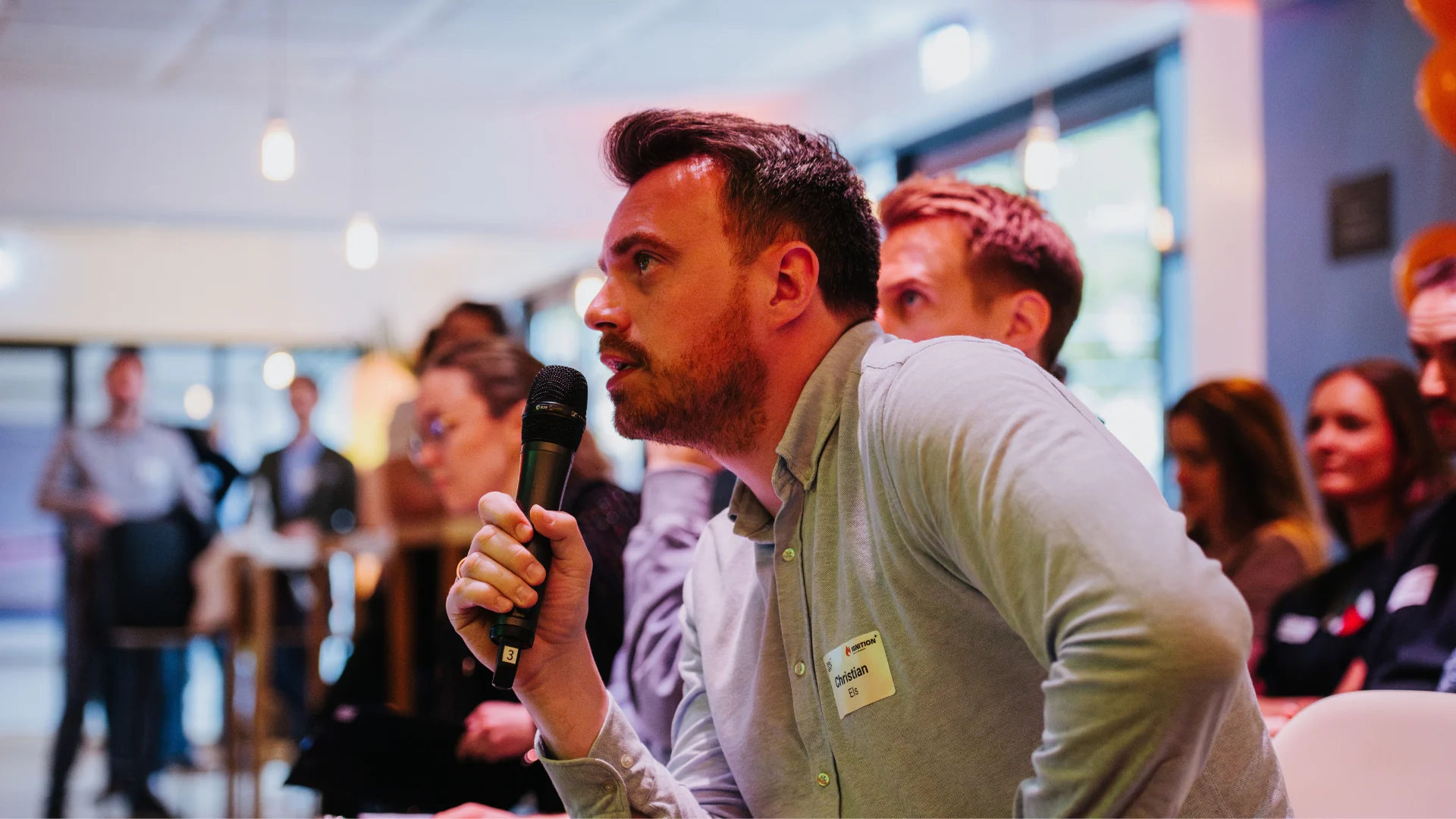 Christian Eis holding a microphone and speaking attentively during an indoor event with blurred audience in the background.